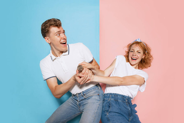 Portrét mladého emotivního páru bojujícího o zpívající mikrofon izolovaný na růžovém modrém pozadí studia. Karaoke bitva. Pojem mládí, emoce, výraz tváře, láska, vztah. Plakát, inzerát - Fotografie, Obrázek