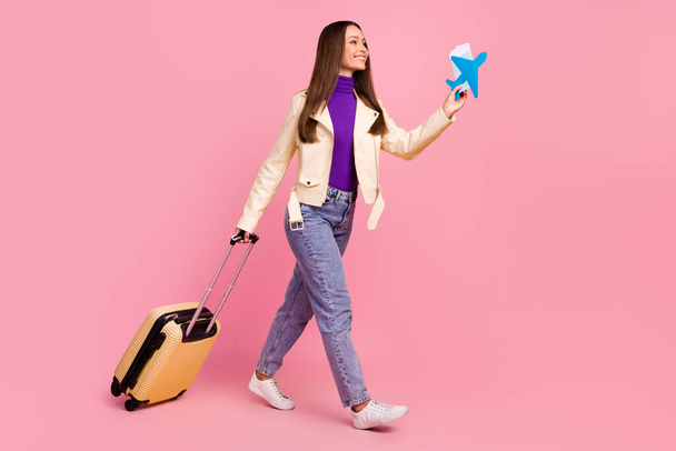 Full length profile photo of cute young lady with valise hold plane look promotion wear jacket jeans boots isolated on pink color background. - 写真・画像