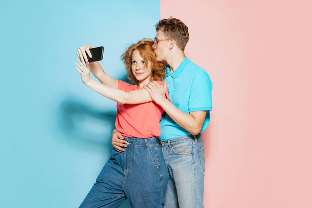 Pembe mavi stüdyo arka planında izole edilmiş telefonla selfie çeken genç çiftin portresi. Öpüşme. Gençlik kavramı, duygular, yüz ifadesi, aşk, ilişki. Poster, reklam - Fotoğraf, Görsel