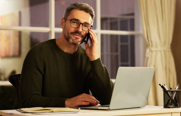 Altijd oproepbaar. Gesneden portret van een knappe volwassen zakenman het maken van een telefoongesprek tijdens het werken op zijn laptop thuis - Foto, afbeelding