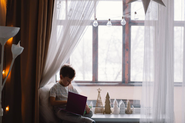 Милий хлопчик читає книжку, що сидить на підвіконні біля вікна. Затишний будинок з декоративними ялинками. Солодкий дім. Зимові канікули спосіб життя
. - Фото, зображення