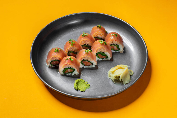Sushi roll stuffed with salmon and lettuce, wrapped in rice, topped with crab meat and tuna sashimi, sprinkled with finely chopped green onions. Sushi stands on a black, round, ceramic plate. The plate stands on a yellow paper background. - Photo, Image