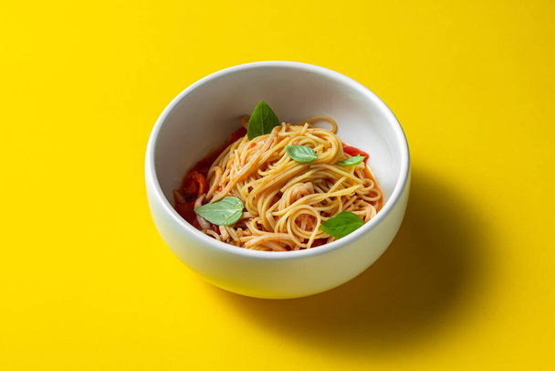 Homemade pasta with basil leaves, tomatoes and tomato sauce. Pasta lies in a light, deep ceramic plate. The plate stands on a yellow paper background. - Foto, Imagen