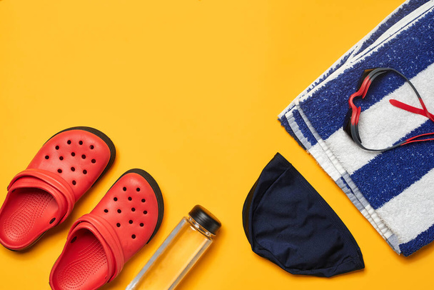 Flat lay summer pool accessories such as swimming cap and goggles, transparent bottle, striped towel and red slippers on a yellow background with copy space. Colorful beach wear. Top view, close-up. - Photo, image