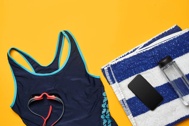 Flat lay summer pool accessories such as blue swimwear, red swimming goggles, striped towel, smartphone and transparent bottle on a yellow background. Colorful beach wear. Copy space, top view, close - Foto, Bild