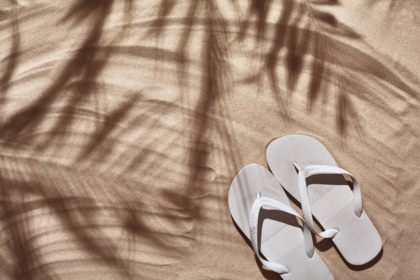 Summer concept with a shadow of a tropical palm tree leaves, copyspace. White thongs are on a perfect beach sand. Summertime lifestyle, objects in flat lay, top view arrangement. - Photo, image