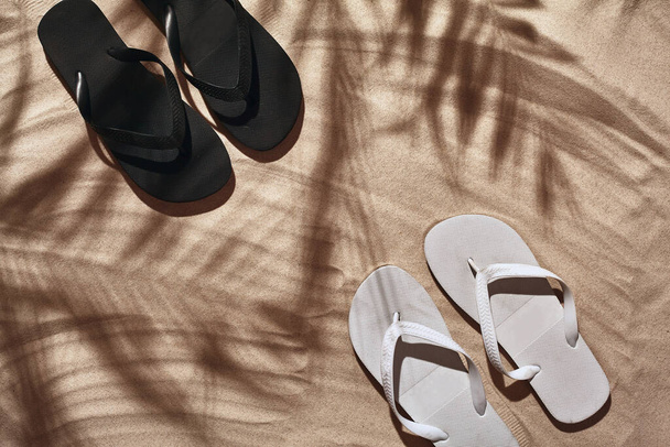 Summer concept with a shadow of a tropical palm tree leaves, copyspace. White and black thongs are on a perfect beach sand. Summertime lifestyle, objects in flat lay, top view arrangement. - Фото, изображение