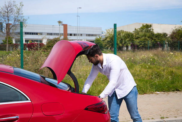 Young, handsome man opens the boot of his red sports car. The man is wealthy. High standard of living and well positioned financially. - Fotografie, Obrázek