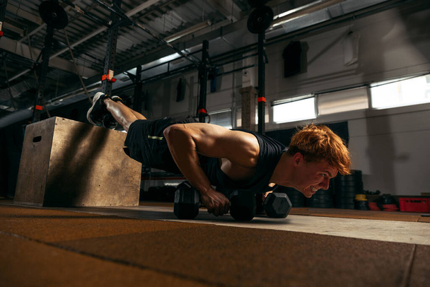 Endurance. Portrait of young sportive red-headed man doing push up exercises with dumbbells isolated on indoor gym background. Concept of sport, health, action, nutrition, youth. Copy space for ad - Foto, Imagem