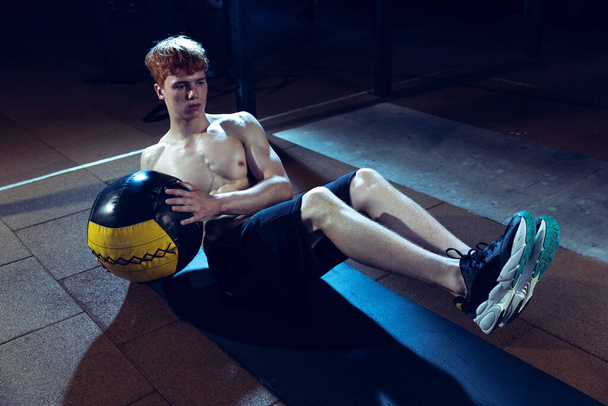 Portrait of young sportive, muscular man doing press exercises with fitness ball isolated over gym background. Concept of sport, health, action, nutrition, youth. Copy space for ad - Photo, image