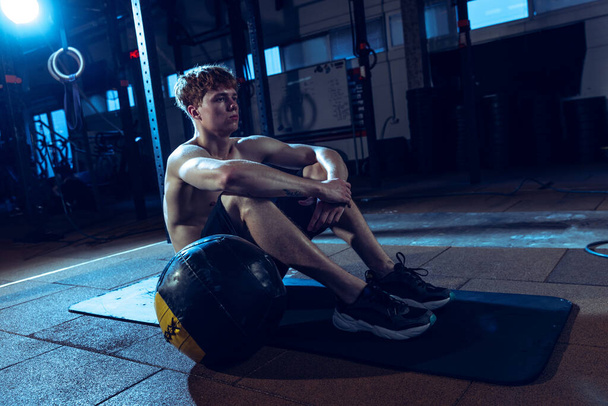 Portrait of young sportive man sitting on fitness rug isolated over gym background. Resting. Workout routine. Concept of sport, health, action, nutrition, youth. Copy space for ad - Foto, afbeelding