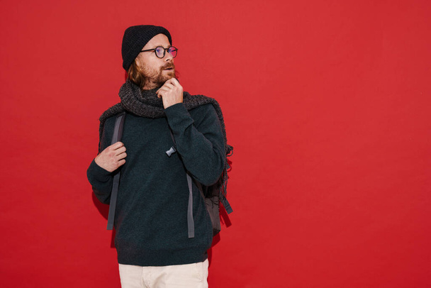 Ginger puzzled man wearing hat and scarf looking aside isolated over red background - Photo, image