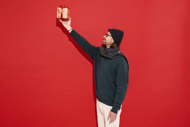 White man in warm clothes and eyeglasses showing gift box isolated over red background - Fotoğraf, Görsel