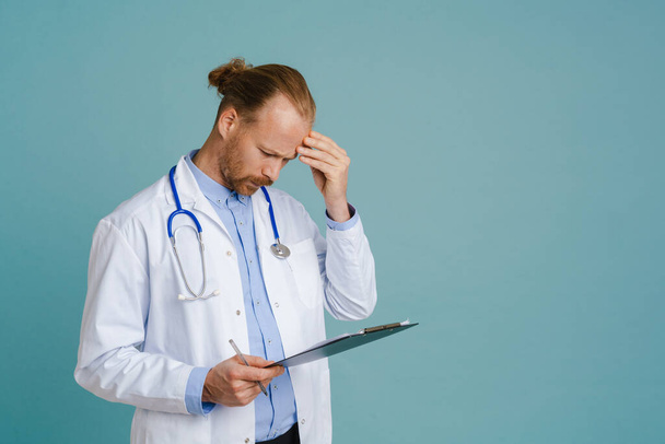 Puzzled male doctor wearing lab coat posing with clipboard isolated over blue background - Zdjęcie, obraz