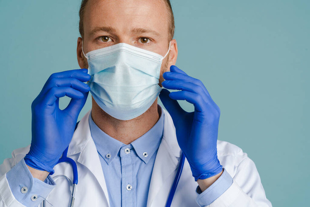 White male doctor posing in face mask and medical gloves isolated over blue background - Photo, Image
