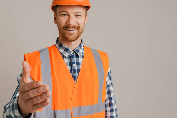 White man worker wearing helmet smiling and stretching hand at camera isolated over grey background - Foto, afbeelding