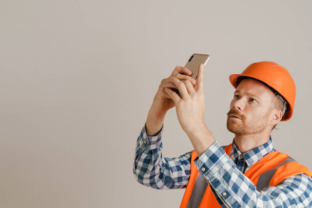 White man worker wearing helmet and vest using mobile phone isolated over grey background - Zdjęcie, obraz