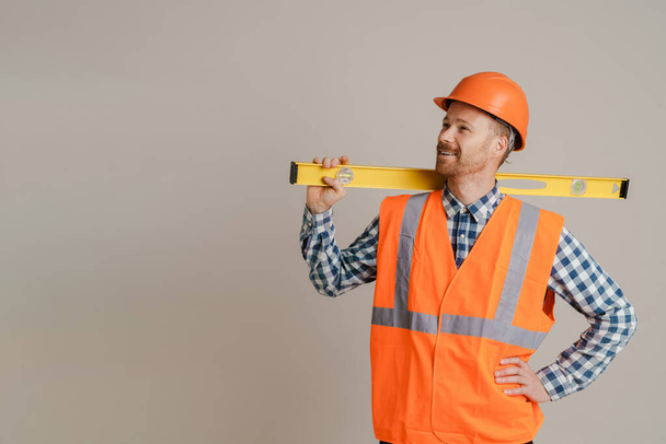 White man worker wearing helmet and vest posing with spirit level tool isolated over grey background - Φωτογραφία, εικόνα