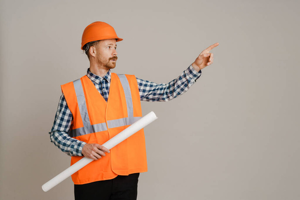 White man builder pointing finger aside while holding drawing isolated over grey background - Фото, зображення
