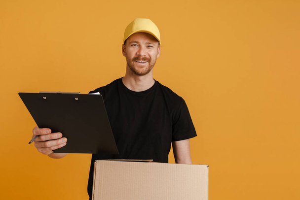 White delivery man in cap posing with cardboard box and clipboard isolated over yellow background - Foto, imagen