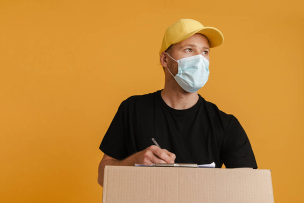 White delivery man in face mask writing down notes on clipboard isolated over yellow background - Foto, afbeelding