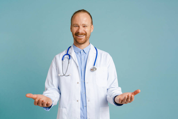 White male doctor wearing lab coat smiling and holding copyspace isolated over blue background - Foto, afbeelding