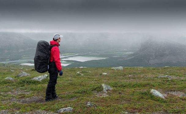 Female hiker in red jacket with heavy backpack overlooking Rapa river valley in arctic landscape of Sarek National Park, Sweden, on a cloudy and rainy day. Hiking in the far north. Rain in the arctic - Photo, image