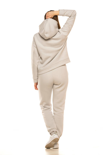 Rear view of a young woman in a gray tracksuit and hood posing to a white background in the studio. - Φωτογραφία, εικόνα