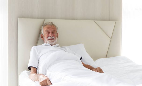 An elderly Caucasian man woke up and smiled. After a good night's sleep, he was resting in bed alone in the bedroom. Healthy concept, health care, old people, medical and elderly retirement. - Foto, immagini