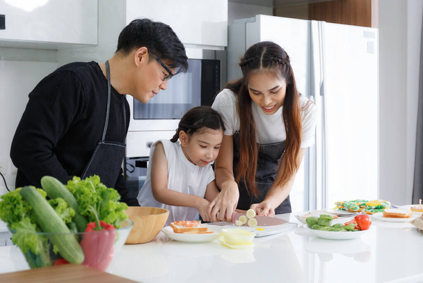 Happy family cooking together in the kitchen. Father, mother, and cute little daughter turn vegetables to make salads. Prepare food in the kitchen at home. Happy family relationship concept Lifestyle. - Фото, изображение