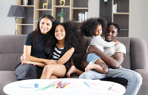 happy multi-ethnic family cuddling on the sofa with cute daughter, couple smiling, looking at the camera, Asian mom and black dad Relax together in the living room at home. family relationship concept - Photo, image