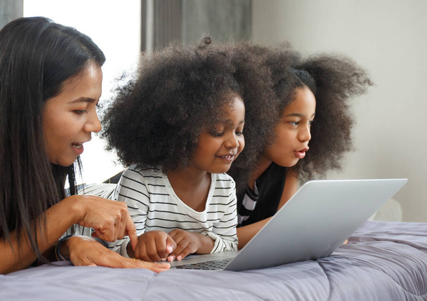 Portret van een Aziatische moeder met een multi-etnische dochter Twee schattige kleine meisjes met laptops op zoek naar studievrienden in bed in de slaapkamer thuis, concept, groei, ontwikkeling, technologie. - Foto, afbeelding
