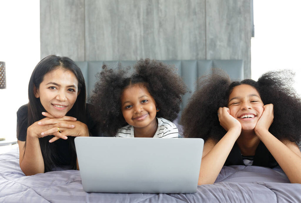 Portret van een Aziatische moeder met een multi-etnische dochter Twee schattige kleine meisjes met laptops op zoek naar studievrienden in bed in de slaapkamer thuis, concept, groei, ontwikkeling, technologie. - Foto, afbeelding