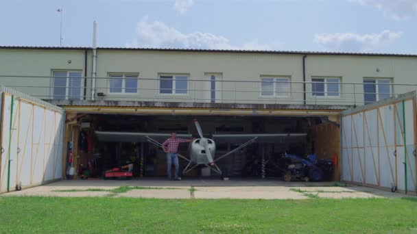 Aviator standing close airplane in hangar small aerodrome countryside. Pilot man posing near ultralight private plane inside repair facility after transportation flight. Aviation business concept. - Materiał filmowy, wideo