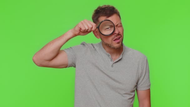 Investigator researcher scientist man holding magnifying glass near face, looking into camera with big zoomed funny eye, searching, analysing. Adult stylish guy on chroma key studio background indoors - Felvétel, videó