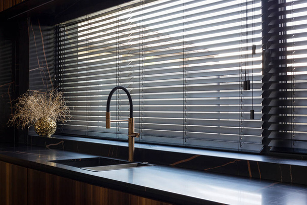 Wooden blinds black color closeup on the window. Bamboo slats 50mm wide. Venetian wood blinds in the kitchen. Black tapes. Sink with copper faucet near the window. Round vase is on the windowsill. - Foto, Imagem