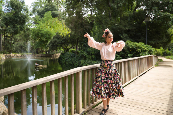Young teenage woman in pink shirt, black skirt with flowers and pink carnations in her hair, dancing flamenco on wooden bridge by a lake. Flamenco concept, dance, art, typical Spanish dance. - Foto, imagen