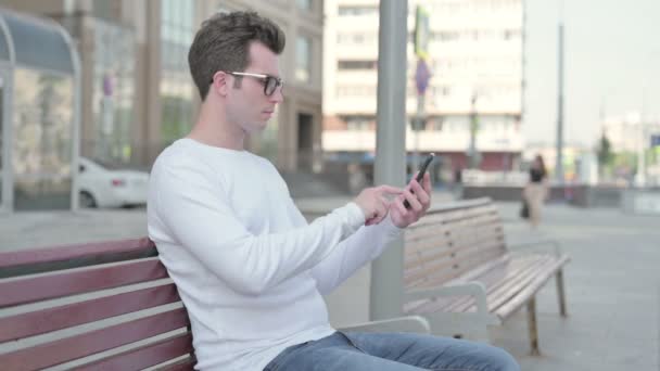 Πλευρική άποψη του Casual Man χρησιμοποιώντας Smartphone, ενώ κάθεται σε εξωτερικούς χώρους για Bench - Πλάνα, βίντεο