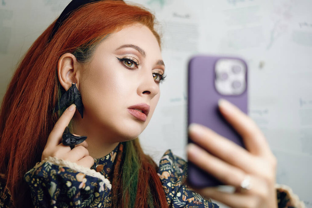 Cute girl takes selfie on front camera of modern smartphone. Female blogger films herself for fashion and makeup blog. Red-haired woman points her finger at her butterfly earrings. Gothic aesthetic. - Foto, afbeelding