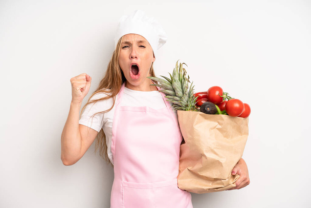pretty caucasian woman shouting aggressively with an angry expression. chef with a market vegetables bag - Foto, afbeelding