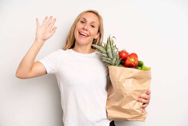 pretty caucasian woman smiling happily, waving hand, welcoming and greeting you. chef with a market vegetables bag - Foto, immagini