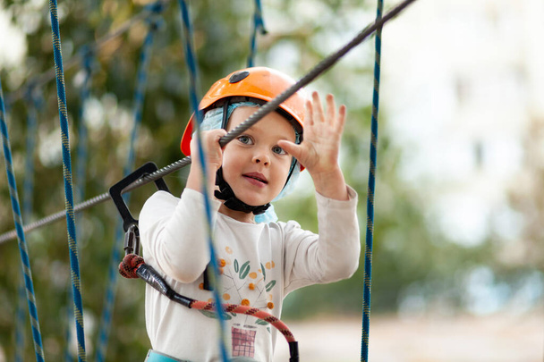 Little girl in helmet climbs ropes in adventure park outdoors. Extreme sport, active leisure on nature. - Zdjęcie, obraz