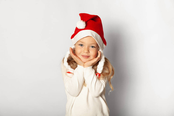 Pienen nätin tytön muotokuva joulupukin hatussa valkoisella taustalla. Hyvää joululomaa, söpö lapsi onnellisuudessa. - Valokuva, kuva