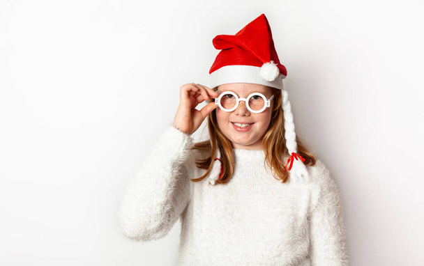 Red-haired girl with freckles and in a santa hat holds hand glasses, isolated on a white background. - Foto, immagini