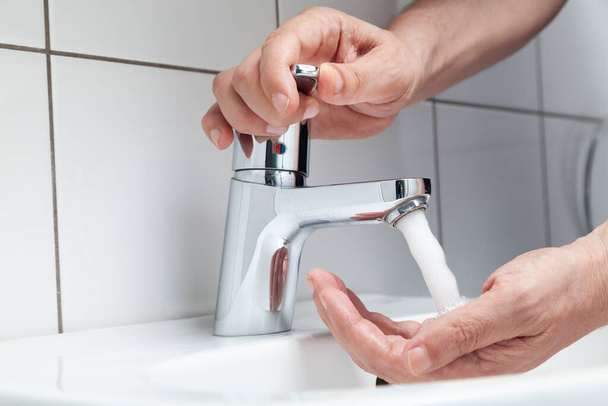 The right hand opens the tap on the lever faucet above the white tiled sink. A jet of water pours on the left hand. White tiles on the wal - Photo, image
