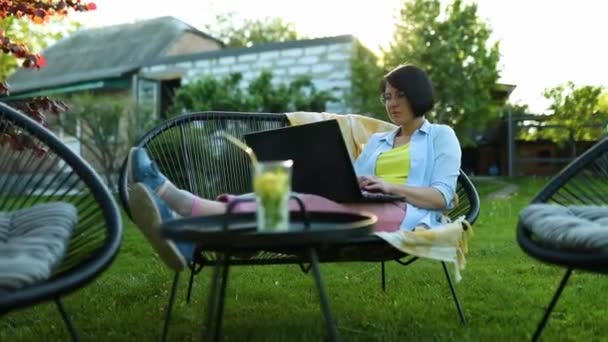 Женщина, печатающая на ноутбуке для удаленной работы, фрилансер, работающая с ноутбуком, проверяющая социальные сети, делающая покупки онлайн, заказывающая доставку дома на открытом дворе в саду - Кадры, видео