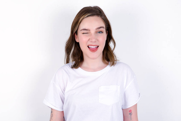 jonge blanke vrouw draagt witte T-shirt over witte achtergrond knipperende ogen met plezier met vrolijke uitdrukking. Gezichtsuitdrukkingen en mensen emoties concept. - Foto, afbeelding