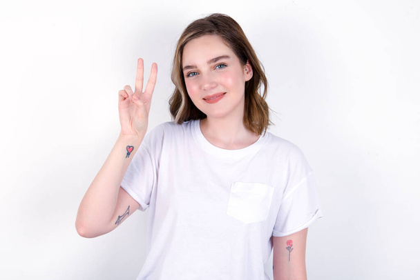 νεαρή καυκάσια γυναίκα φορώντας λευκό T-shirt πάνω από λευκό φόντο δείχνει και δείχνει προς τα πάνω με τα δάχτυλα νούμερο δύο, ενώ χαμογελά αυτοπεποίθηση και χαρούμενος. - Φωτογραφία, εικόνα