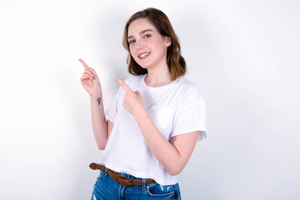 молодая белая женщина в белой футболке на белом фоне с положительным выражением лица, указывает пальцем на пустое место для копирования рекламного текста или рекламы. - Фото, изображение