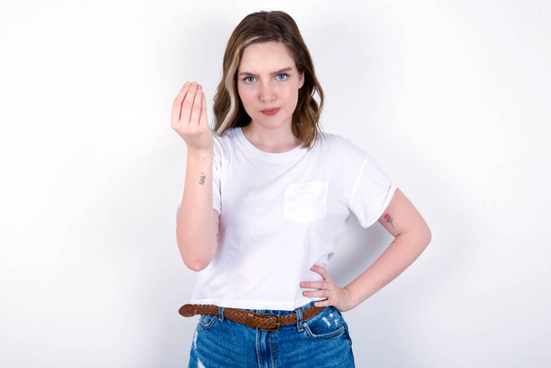 What the hell are you talking about. Shot of frustrated young caucasian woman wearing white T-shirt over white background gesturing with raised hand doing Italian gesture, frowning, being displeased and confused with dumb question. - Fotó, kép